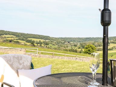 A patio table with a wine glass and cushions on a chair overlooking green rolling hills and fields at Farm View Lodge in Painswick