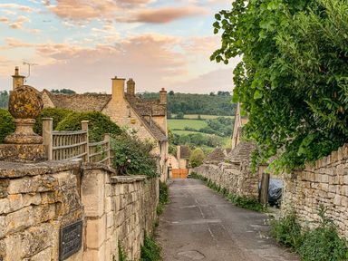 A narrow street with stone walls and cottages with a green hill in the background at Farm View Lodge in Painswick