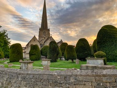 A church with a tall spire behind a garden with trimmed bushes and stone structures at Farm View Lodge in Painswick