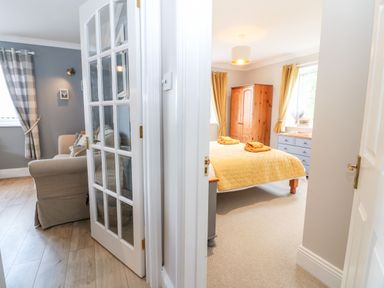 A view showing a living room with armchairs and checkered curtains and a bedroom with a bed with a yellow quilt and wooden furniture at Farm View Lodge in Painswick