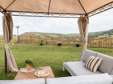 An outdoor seating area under a canopy with a sofa and coffee table overlooking a grassy yard with a wooden fence and hills in the background at Farm View Lodge in Painswick