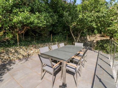 An outdoor patio with a dining table and eight chairs surrounded by trees and stone walls at Westwinds in Tenby