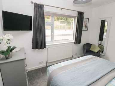 A bedroom with a bed a grey chest of drawers a black mounted TV a chair and a window with dark curtains at Westwinds in Tenby
