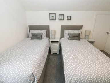 A bedroom with two single beds side by side with grey and white bedding a nightstand with a lamp between the beds and three small wall shelves above at Westwinds in Tenby