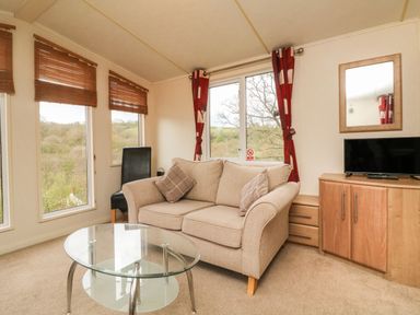A living room with a beige sofa, glass coffee table, black chair, and wooden cabinet with a TV at Exe Valley Lodge in Bampton Devon