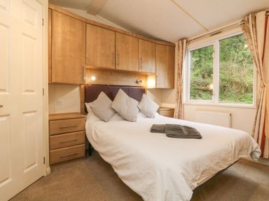 A bedroom with a bed covered in white bedding and three pillows under built in wooden cabinets next to a window at Exe Valley Lodge in Bampton Devon