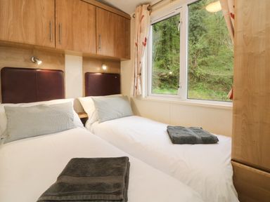 A bedroom with two single beds, pillows, towels on beds, wooden overhead cabinets, and a window showing greenery at Exe Valley Lodge in Bampton Devon