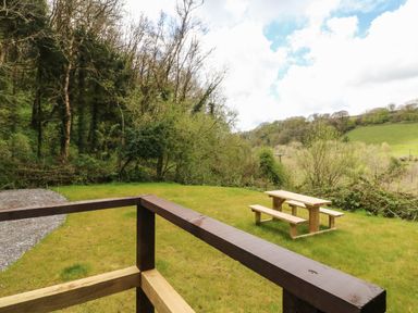 A garden area with wooden picnic table and benches surrounded by trees and grassy hill at Exe Valley Lodge in Bampton Devon