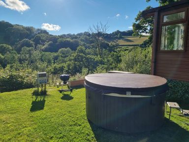 An outdoor area with a hot tub with cover a small barbecue grill a power control unit and a chair near a wooden cabin with trees in the background at Exe Valley Lodge in Bampton Devon