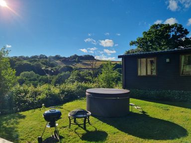 An outdoor area with a hot tub a barbecue grill and a fire pit beside a dark wooden building at Exe Valley Lodge in Bampton Devon