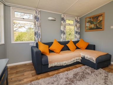 A sitting room with a dark blue sofa with orange cushions and beige throws next to windows with floral curtains at Valley Lodge in Bampton Devon