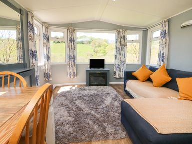 A living room with a wooden dining table and chairs a gray carpet and blue sofas with orange cushions at Valley Lodge in Bampton Devon