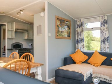 A dining table with wooden chairs and a kitchen area with stove beside a living area with a couch and orange cushions at Valley Lodge in Bampton Devon