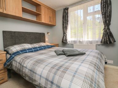 A bedroom with a double bed with plaid bedding two folded towels on the bed wooden bedside tables and wall cabinets a window with curtains at Valley Lodge in Bampton Devon