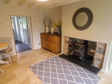 A room with a wood stove in a stone fireplace a circular mirror above a wooden sideboard with a lamp and flowers a gray patterned rug and a wooden chair at Sea View Newport Pembrokeshire
