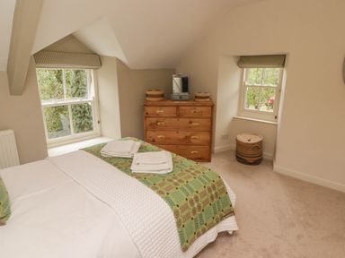 A bedroom with a bed covered with a green patterned blanket a wooden dresser two windows with striped valances and a woven basket at Sea View in Newport Pembrokeshire