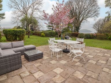 A backyard patio with outdoor seating including a gray sofa, glass-top table, white metal table with four chairs, and a grassy area with trees and shrubs at Sea View in Newport Pembrokeshire