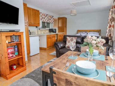 A kitchen dining and living area with wooden cabinets a dining table set with bowls and wine glasses a brown leather sofa and a bed in the background at The Pool House in Crediton