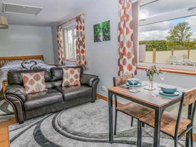 A room with a brown leather sofa and two cushions a wooden dining table set for two and a bed near windows at The Pool House in Crediton
