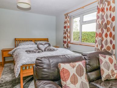 A bedroom with a wooden bed grey bedding and towel swans next to a window with leaf patterned curtains and a leather sofa with matching cushions at The Pool House in Crediton