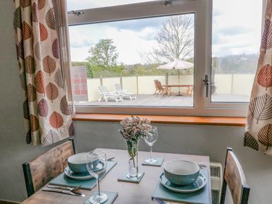 A dining table set with bowls wine glasses and cutlery next to a window overlooking outdoor patio furniture and sun loungers at The Pool House in Crediton