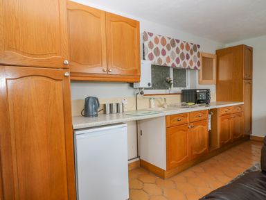 A kitchen with wooden cabinets a white mini fridge a kettle a microwave and a window with a patterned blind at The Pool House in Crediton