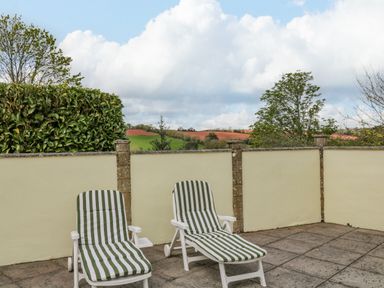 An outdoor patio with two striped lounge chairs and a low wall overlooking trees and fields at The Pool House in Crediton