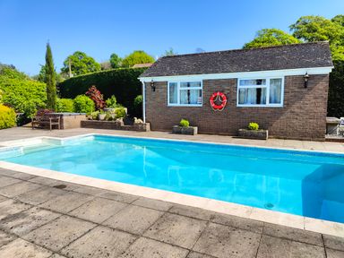 An outdoor swimming pool with a bench and greenery next to a brick building at The Pool House in Crediton