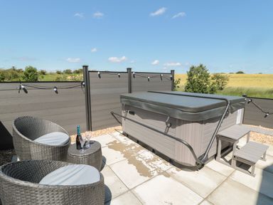 An outdoor patio with a hot tub covered with a lid a small table with a bottle and glasses and two round chairs near a grey fence at Dreams & Streams Bockenfield Country Park near West Thirston
