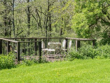 A wooden deck with white metal chairs and a table surrounded by green trees and grass at Little Barton in Stogumber near Watchet