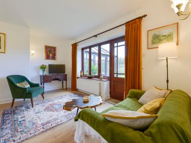 A living room with a green sofa, wooden coffee table, armchair, desk with TV, and large window with orange curtains at Little Barton in Stogumber near Watchet