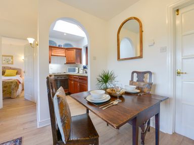A dining area with a wooden table set for two next to an archway leading to a kitchen with wooden cabinets and a bedroom visible through an open door at Little Barton in Stogumber near Watchet