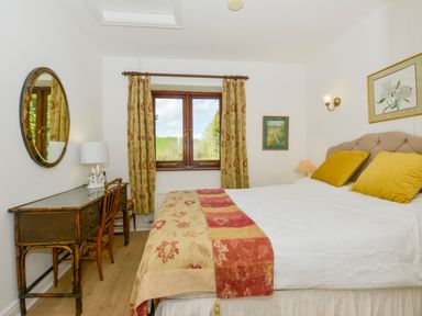 A bedroom with a double bed and yellow pillows a desk with a chair and a lamp by a window with curtains at Little Barton in Stogumber near Watchet