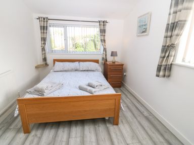 A bedroom with a wooden bed with gray patterned bedding two rolled towels on the bed a wooden nightstand with a lamp a window with plaid curtains and light gray wood flooring at Beach Reach in Valley