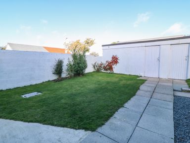 A backyard with green grass shrubs a white wall and a concrete path leading to white doors at Beach Reach in Valley