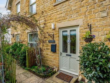 The exterior door of a stone cottage with plants a wooden fence and hanging flower baskets at Jasmine Cottage in Mickleton Gloucestershire