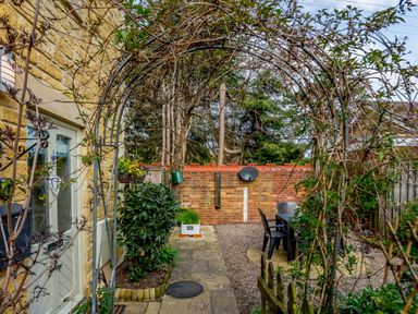 A garden area with a stone pathway a metal arch with climbing plants outdoor table and chairs near a brick wall at Jasmine Cottage in Mickleton Gloucestershire