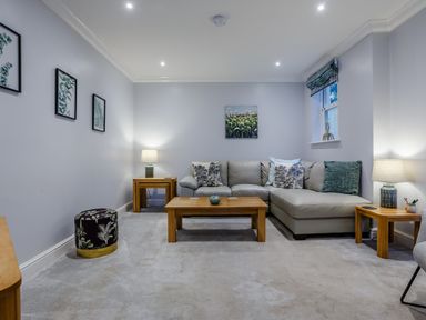 A living room with a grey sofa wooden coffee and side tables lamps and framed pictures on walls at Jasmine Cottage Mickleton Gloucestershire