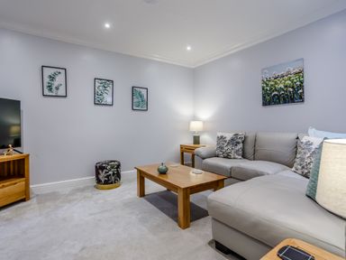 A living room with a grey sectional sofa wooden coffee table wooden TV stand and framed botanical prints on walls at Jasmine Cottage in Mickleton Gloucestershire