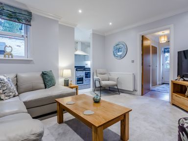 A living room with a gray sofa a wooden coffee table and an armchair with a view into a kitchen at Jasmine Cottage in Mickleton Gloucestershire