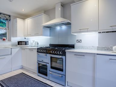 A kitchen with white cabinets a blue oven stove a microwave toaster kettle and a gray rug on the floor at Jasmine Cottage in Mickleton Gloucestershire