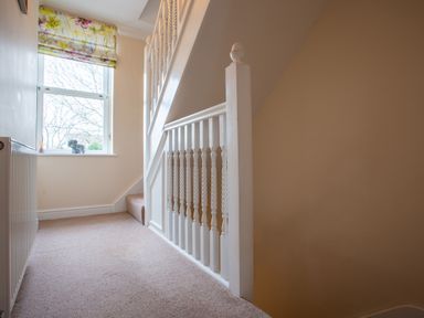 A hallway with carpeted floor a white staircase railing a window with floral curtains and a small decorative object on the windowsill at Jasmine Cottage in Mickleton Gloucestershire