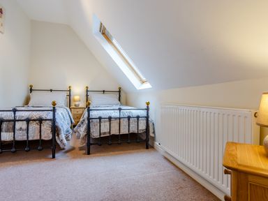 A bedroom with two single beds a wooden nightstand with a lamp a slanted skylight window and a radiator at Jasmine Cottage in Mickleton Gloucestershire