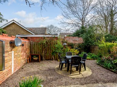 A garden with a round table and four chairs on a gravel area surrounded by plants and brick walls at Jasmine Cottage in Mickleton Gloucestershire