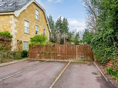 A paved driveway with three parking spaces and a wooden fence next to a stone house surrounded by greenery at Jasmine Cottage in Mickleton Gloucestershire