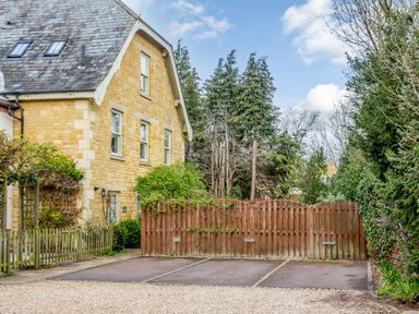 A stone house with a slate roof next to a wooden fence and parking spaces surrounded by trees at Jasmine Cottage in Mickleton Gloucestershire