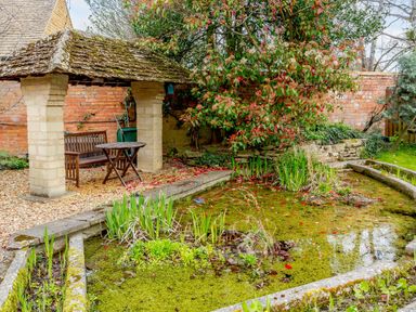 A garden area with a pond a covered bench and a wooden table at Jasmine Cottage in Mickleton Gloucestershire