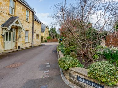 A driveway with stone houses on the left and a garden with plants and a tree on the right at Mickleton House in Mickleton Gloucestershire