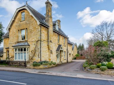 A stone house with windows and a driveway next to a road with plants and trees at Jasmine Cottage Mickleton Gloucestershire