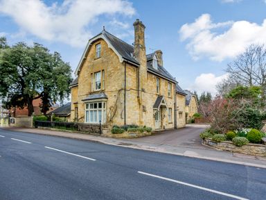 A stone house with multiple chimneys near a road with shrubs and trees at Jasmine Cottage in Mickleton Gloucestershire